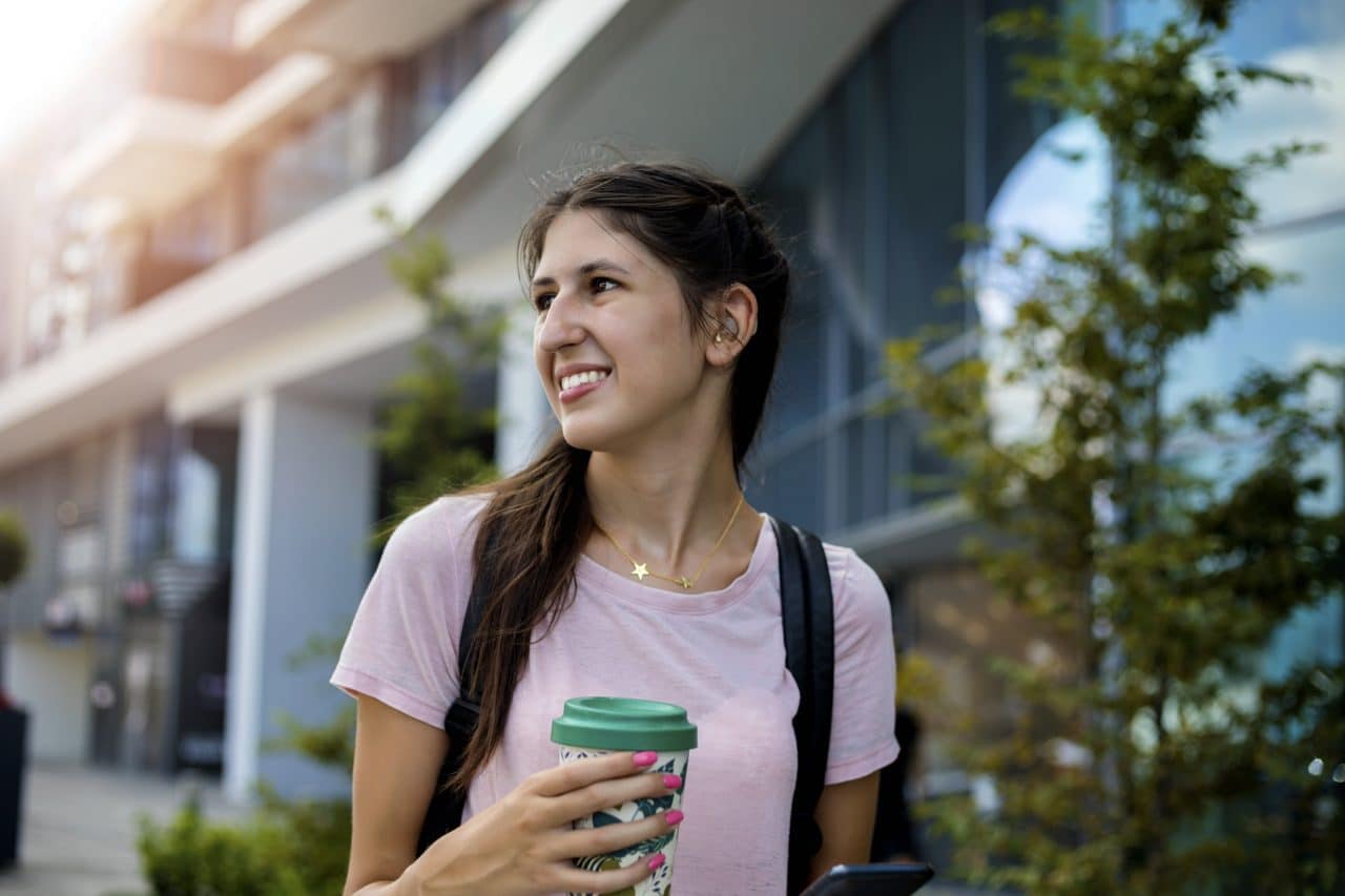 a woman wearing hearing aids walks past a modern building while holding a reusable coffee cup