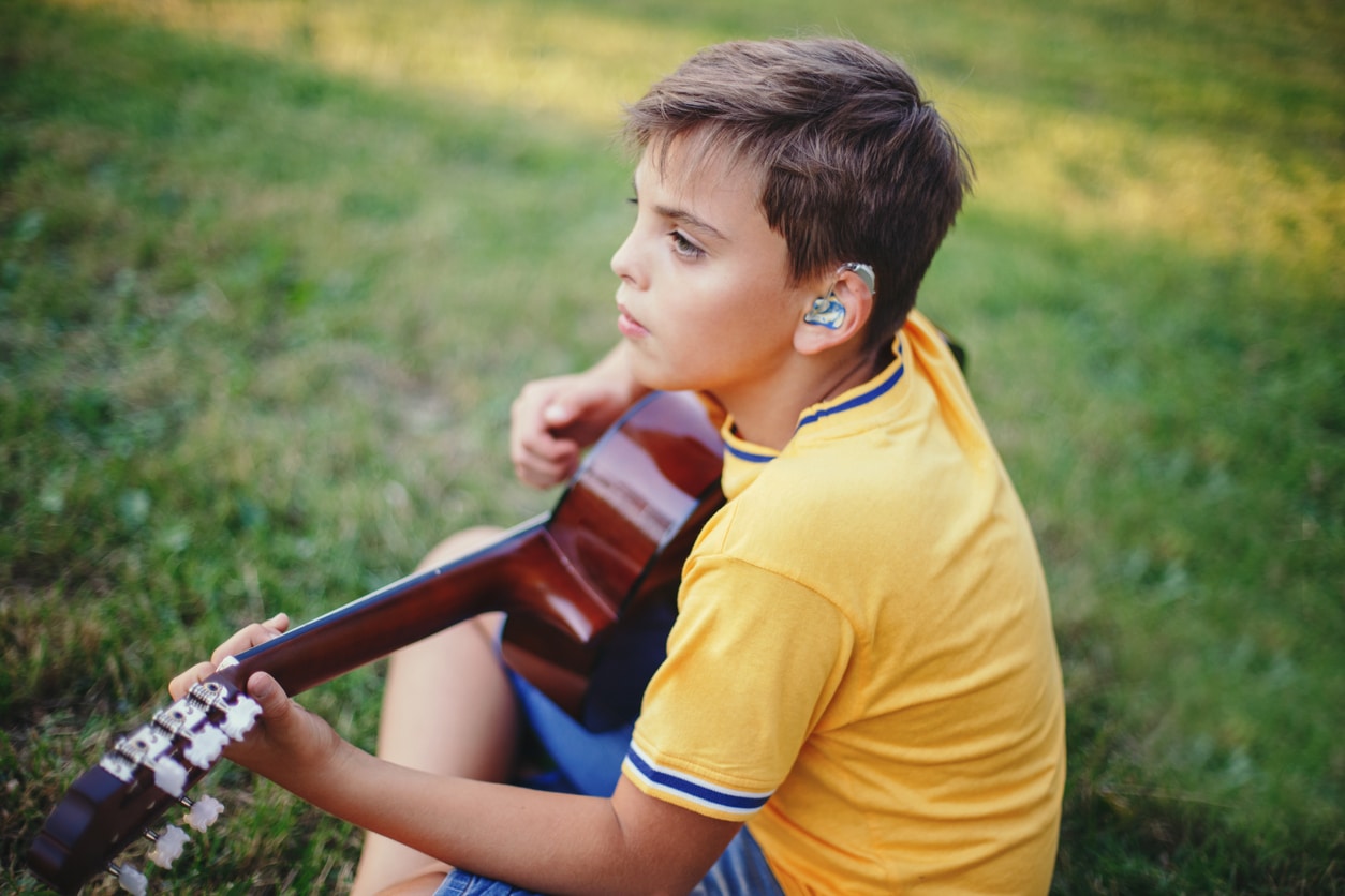 Young boy holding a guitar wearing a hearing aid sitting in grass